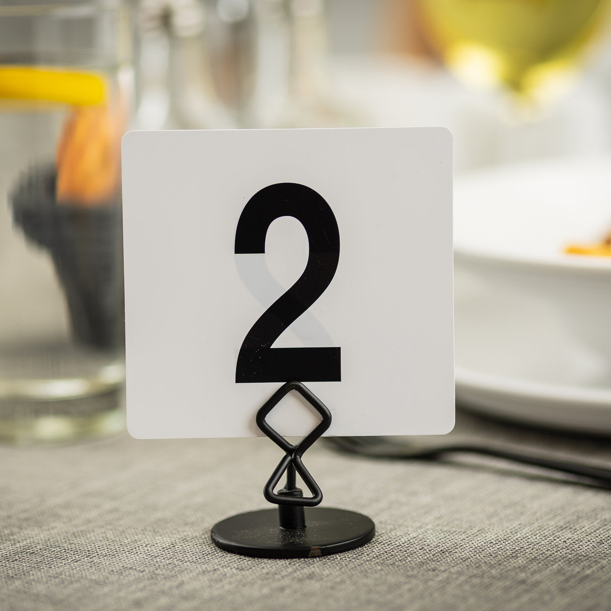 A white square card with a bold black number 2 is held upright by a black powder coated metal Number/Card Holder Stand on a gray tablecloth. In the blurred background, there’s a glass of water with lemon, a white plate, and other dining items.
