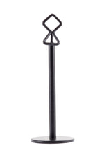A black powder coated metal paper towel holder with a round flat base, vertical cylindrical rod, and a unique diamond-shaped loop at the top for easy carrying. The minimalist, modern design resembles a TableCraft number/card holder stand. Photographed on white.