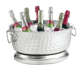 A large, hammered stainless steel beverage tub from the Bali Collection chills nine bottles of sparkling wine with foil-wrapped tops in silver, gold, green, and pink. The double wall beverage tub has side handles and sits on a matching silver base.