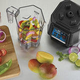 A TORQ 2.0 Series Blender with its copolyester container filled with sliced bell peppers, onions, and cilantro sits next to the motor base. Beside it, cut mangoes, tomatoes, lime, and more ingredients rest on a cutting board on a light surface.