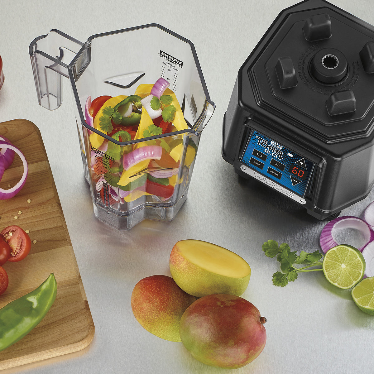 A disassembled TORQ 2.0 Series Blender sits on the counter. Its copolyester container holds sliced onions, peppers, and herbs, while mangoes, a lime, and cilantro rest nearby. The black base features digital controls and an illuminated time readout.