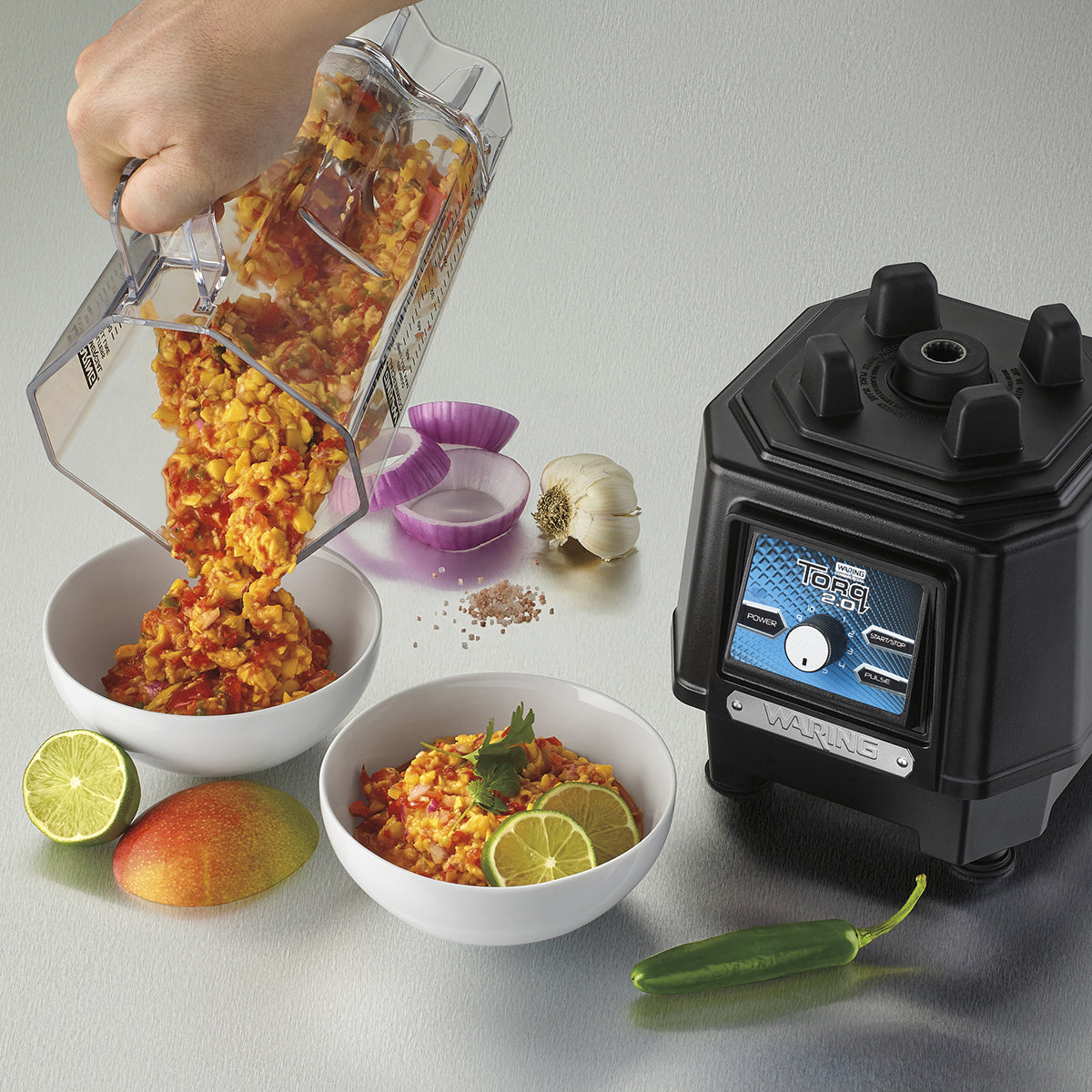 A hand pours a chunky orange and red salsa from a TORQ 2.0 Series Blender with a copolyester container into a white bowl. Another cilantro-garnished salsa sits nearby, surrounded by lime, mango, green chili, garlic, salt, and sliced red onion.
