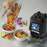 A hand pours a chunky orange and red mixture from a TORQ 2.0 Series Blender copolyester container into a white bowl. Another garnished bowl sits beside ingredients like mango, lime halves, jalapeño, onion, garlic, and salt; the blender base is on the right.