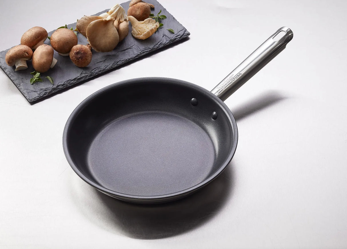 A Vulcano Fry Pan with a stick resistant finish and 18/10 stainless steel handle sits on a light countertop. Next to it, a slate tray displays neatly arranged brown and white oyster mushrooms with fresh green herb sprigs.