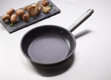 A Vulcano Fry Pan with a stick resistant finish and 18/10 stainless steel handle sits on a light countertop. Next to it, a slate tray displays neatly arranged brown and white oyster mushrooms with fresh green herb sprigs.