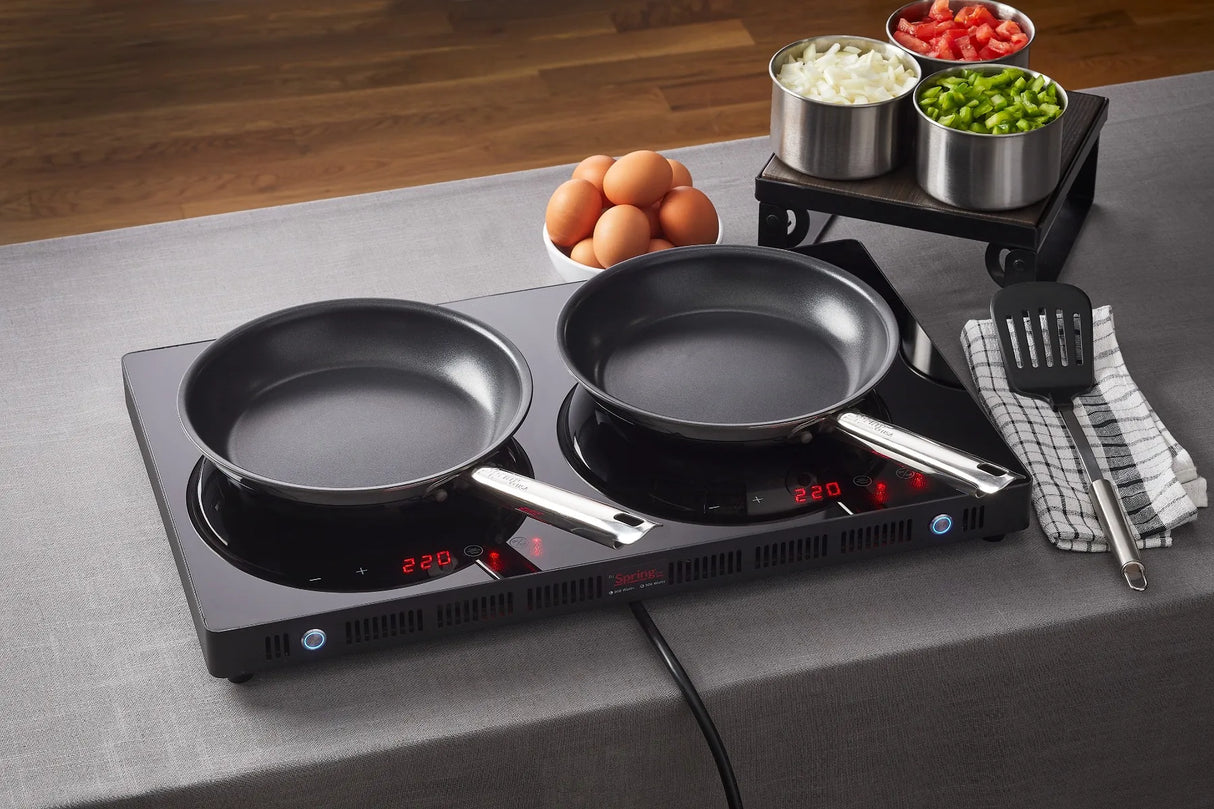 Two black Vulcano Fry Pans with a stick resistant finish sit on a double-burner induction cooktop set to 220°C. Nearby are a striped towel with spatula, brown eggs, and three metal bowls of chopped onions, tomatoes, and green peppers on a gray tablecloth.