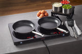 Two black Vulcano Fry Pans with a stick resistant finish sit on a double-burner induction cooktop set to 220°C. Nearby are a striped towel with spatula, brown eggs, and three metal bowls of chopped onions, tomatoes, and green peppers on a gray tablecloth.