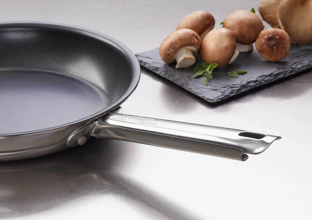 A close-up of the Vulcano Fry Pan with a stick resistant finish sits on a shiny metal countertop. Next to it, mushrooms and green herbs rest on a rectangular black slate plate in the background, perfect for induction cooking.