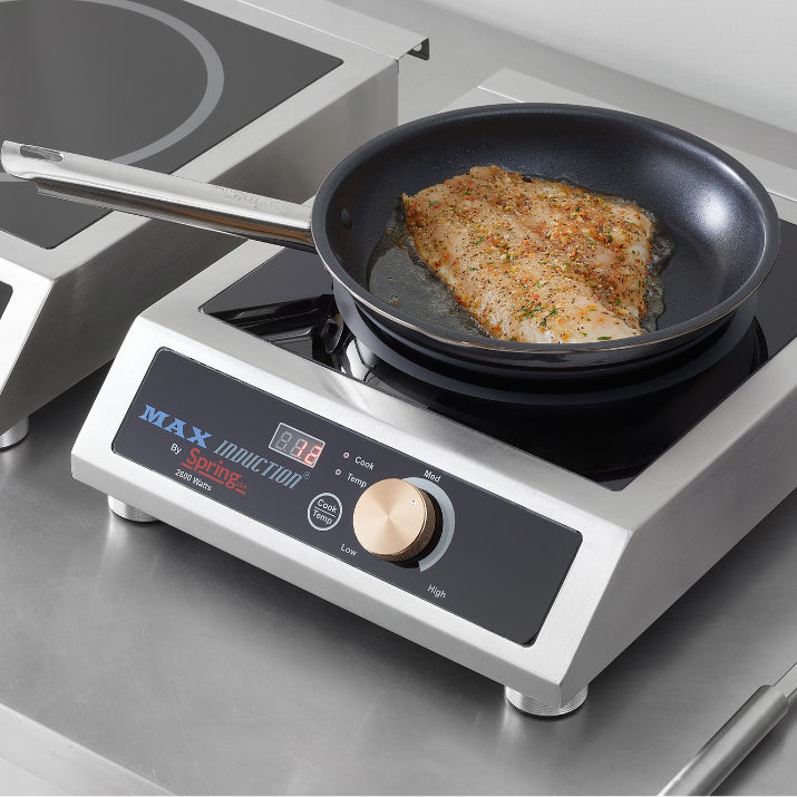 A Vulcano Fry Pan with a seasoned fish fillet cooks on an induction cooktop. The display reads “98” and the control knob is set to medium. The pan’s stick resistant finish ensures easy release as it sits atop a stainless steel counter.