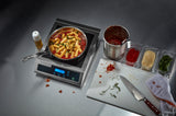 A pan of rigatoni pasta in red sauce with herbs cooks on a single hob Induction Range. Nearby, oil, cheese, tomatoes, basil, chopped herbs, and a stained towel are spread across the gray countertop.