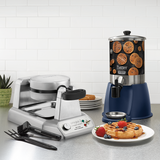 A stainless steel Belgian waffle maker with a waffle inside sits next to a 2 gallon batter dispenser labeled with a waffle image. Plates of golden waffles topped with strawberries, blueberries, and syrup sit in front. A fork, knife, and plant are also visible on the counter.