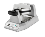 A silver, stainless steel Waring WBW300XCE Double Belgian Waffle Maker with a black handle and base. It features indicator lights, a rotary knob on the front, a deep drip tray below, and is designed for international use. The waffle plates are clamped shut.