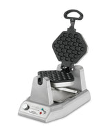 A silver Waring WBW300XCE bubble waffle maker with a hexagonal, honeycomb-patterned nonstick plate is open, revealing both plates. The base has a dial, indicator lights, and a drip tray, making it perfect for international use.