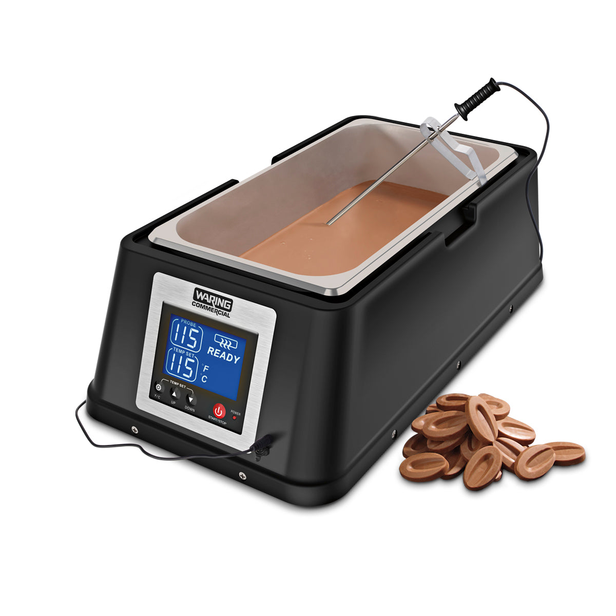 A black Waring chocolate melter with a stainless steel pan and digital display showing “115°F” and “READY” holds melted chocolate. A metal spatula rests inside, while oval chocolate pieces are scattered on the countertop beside the machine.