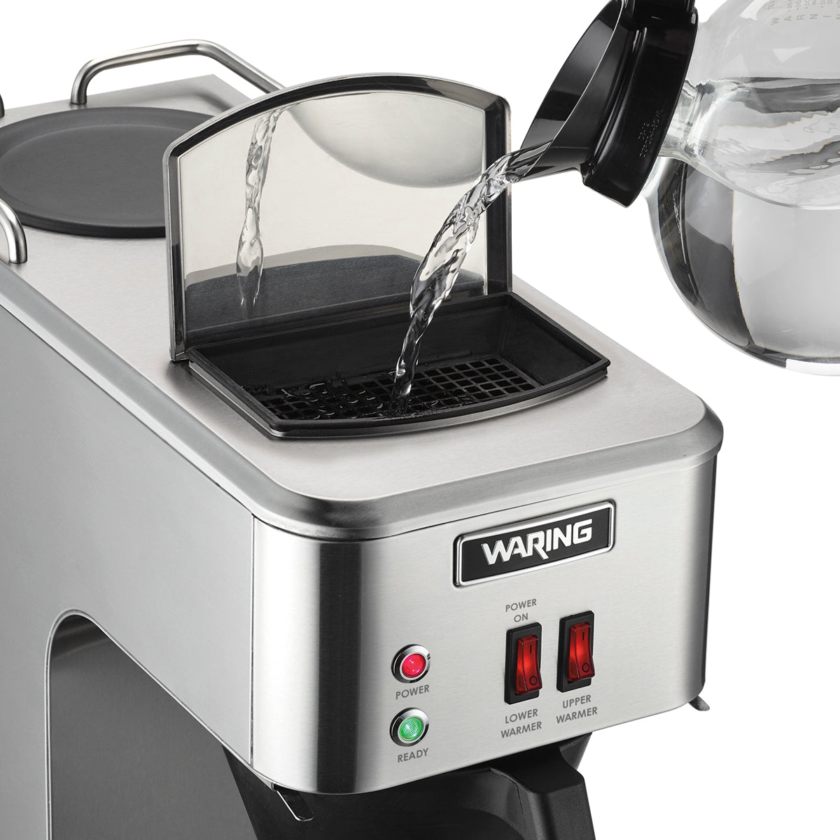 A hand pours water from a clear glass pitcher into the open water reservoir of a commercial coffee maker. The stainless steel pourover coffee brewer features indicator lights and switches labeled “Lower Warmer” and “Upper Warmer” on the front panel.