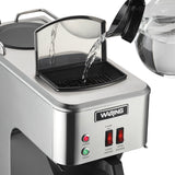 A hand pours water from a clear glass pitcher into the open water reservoir of a commercial coffee maker. The stainless steel pourover coffee brewer features indicator lights and switches labeled “Lower Warmer” and “Upper Warmer” on the front panel.
