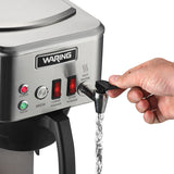 A hand dispenses hot water from a stainless steel Waring commercial coffee machine. The front panel features buttons labeled Power, Brew, Lower Warmer, and Upper Warmer with indicator lights, as water pours from the hot water spout into an unseen container.