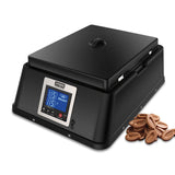 A sleek, modern black countertop chocolate tempering machine with a digital display shows temperature and status. The device features a closed lid, stainless steel accents, and sits beside several oval-shaped pieces of milk chocolate.