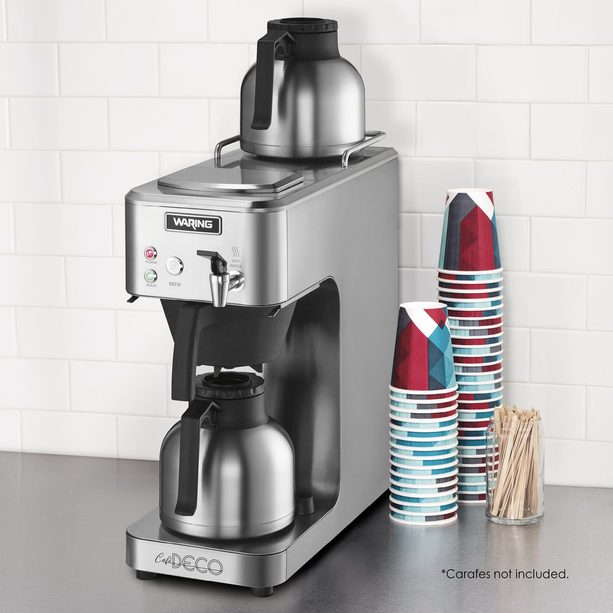 A stainless steel thermal coffee brewer with two metal carafes, one above and one below. Café deco details include stacks of colorful paper cups, a container of wooden stir sticks, and a white tiled wall. Text notes: Carafes not included.