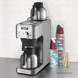 A stainless steel thermal coffee brewer with two metal carafes, one above and one below. Café deco details include stacks of colorful paper cups, a container of wooden stir sticks, and a white tiled wall. Text notes: Carafes not included.