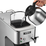 A hand pours water from a metal carafe into the open reservoir of a stainless steel commercial coffee brewer. The machine features labeled buttons for power and brew, a “hot water” spout, and a small screen covering the reservoir opening.