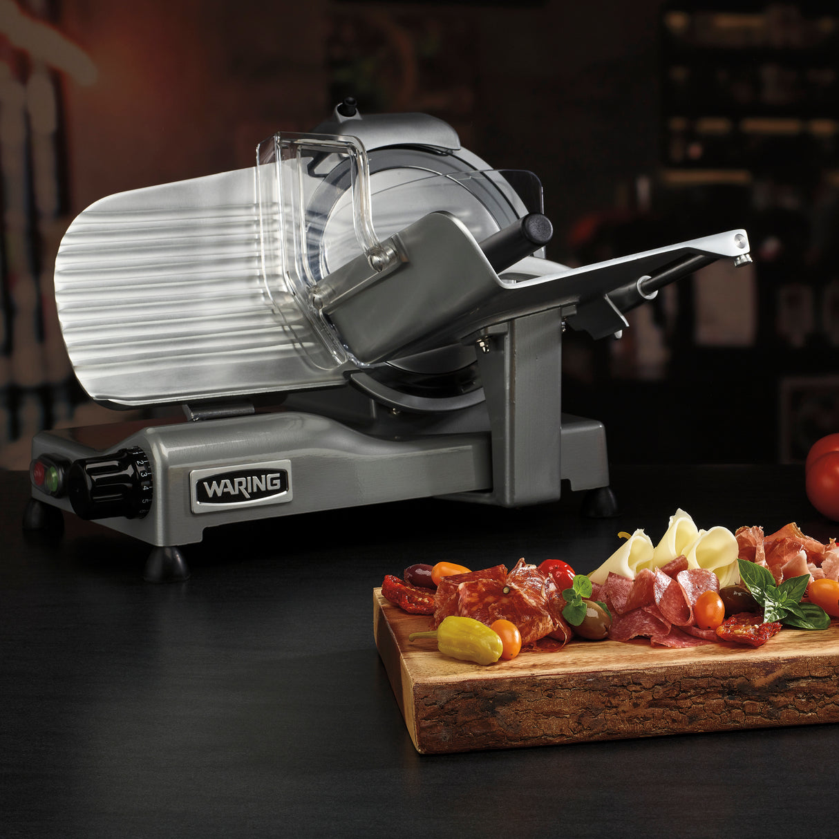 A silver Waring commercial food slicer sits on a dark counter. In front, a wooden board displays sliced cured meats, cheeses, grapes, yellow cherry tomatoes, and herbs. The dimly lit background reveals blurred shelves and kitchen items.