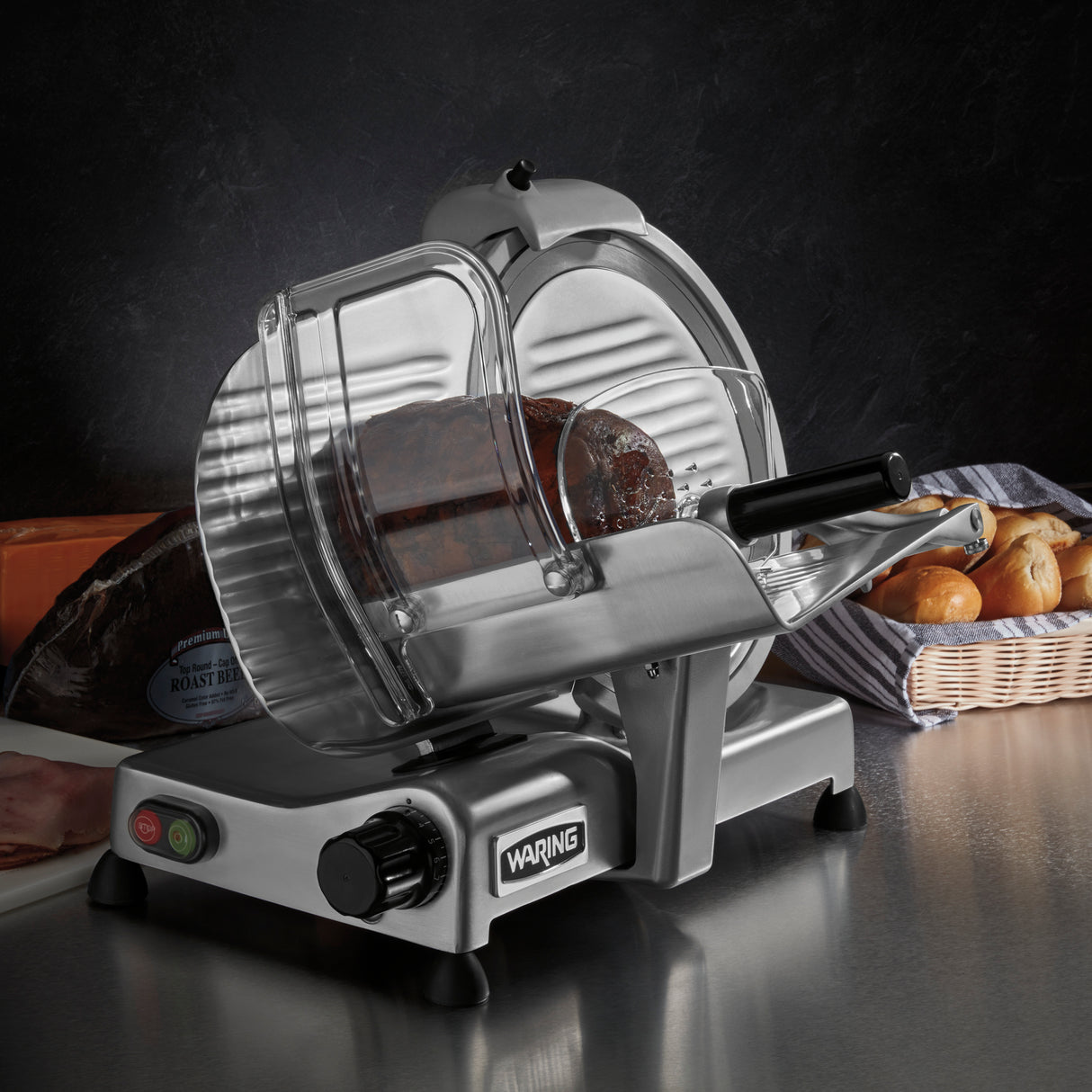 A heavy duty Commercial Food Slicer with a 12" tempered chrome blade slices a roast beef loaf on the counter. The machine features a clear guard, black handle, and control knob. Bread rolls in a basket and wrapped roast beef sit in the dark background.