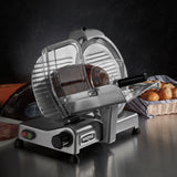 A heavy duty Commercial Food Slicer with a 12" tempered chrome blade slices a roast beef loaf on the counter. The machine features a clear guard, black handle, and control knob. Bread rolls in a basket and wrapped roast beef sit in the dark background.