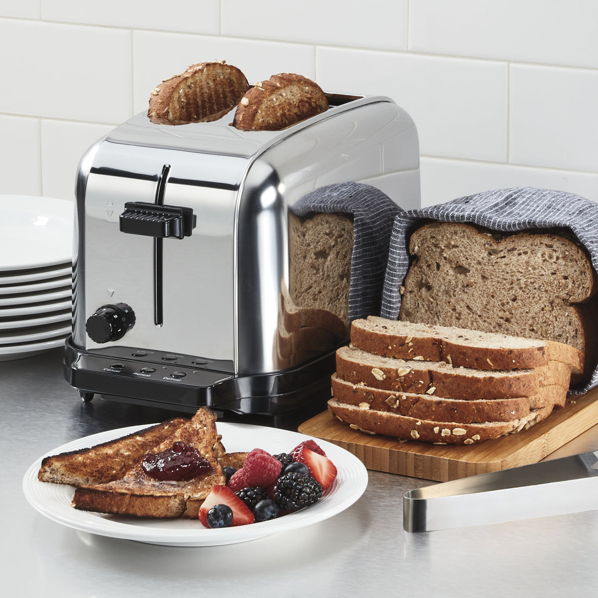 A shiny stainless steel toaster with extra-high lift and browning controls toasts two slices of brown bread on a kitchen counter. Nearby are a loaf of sliced bread, a blue-striped towel, toast with jam, fresh berries, and stacked white plates.
