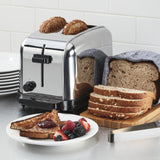 A shiny stainless steel toaster with extra-high lift and browning controls toasts two slices of brown bread on a kitchen counter. Nearby are a loaf of sliced bread, a blue-striped towel, toast with jam, fresh berries, and stacked white plates.