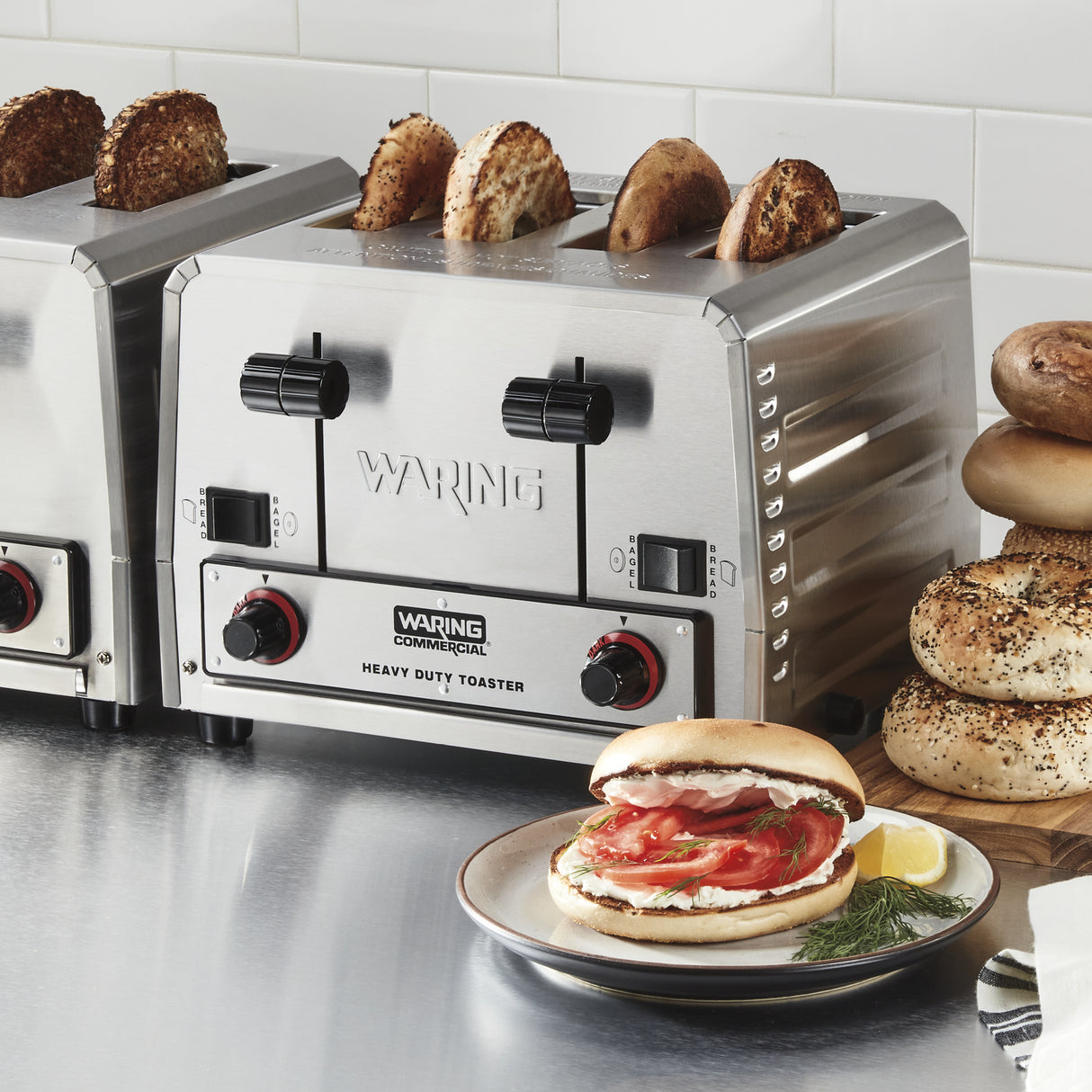 A stainless steel heavy-duty bread toaster with four slots is toasting bagel halves. Nearby, stacked bagels and a plate with a cream cheese, tomato, and dill sandwich sit on a stainless steel countertop against a white tile backsplash.