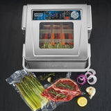 A Waring vacuum packaging machine sits on a dark countertop with its lid open, revealing bags of vegetables inside. In front are vacuum-sealed asparagus and steak with herbs, plus cut onions, garlic, rosemary, pepper, and sliced lemon for garnish.