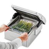A person places a vacuum-sealed bag of green onions into a Waring countertop Vacuum Packaging Machine with the lid open, preparing to seal the bag. The stainless steel machine sits on a white background.