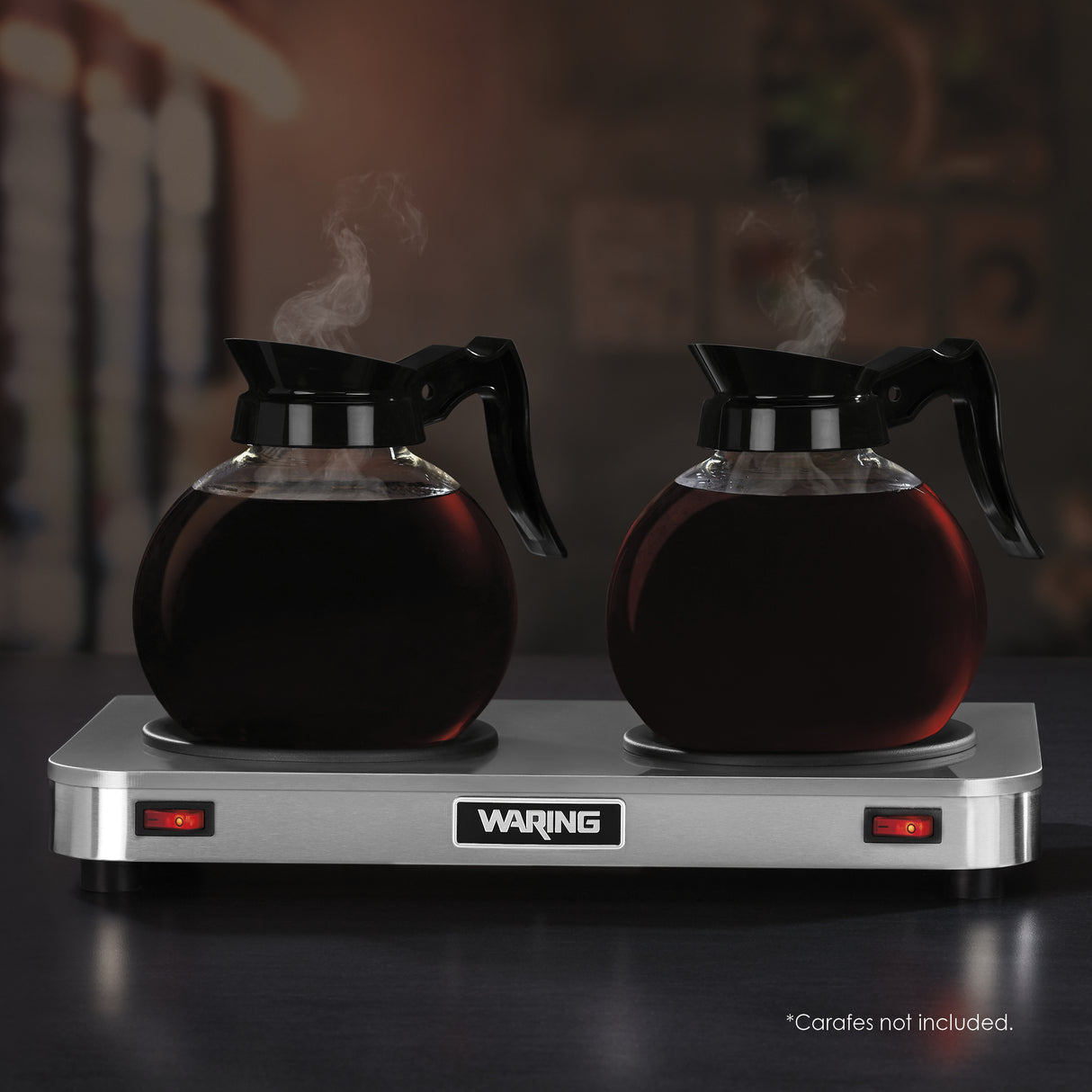 Two glass coffee carafes with black handles sit on a stainless steel double burner coffee warmer. Both carafes are filled with steaming coffee. The stainless steel hot plate has red indicator lights. In the corner, small text reads: “Carafes not included.”.