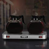 Two glass coffee carafes with black handles sit on a stainless steel double burner coffee warmer. Both carafes are filled with steaming coffee. The stainless steel hot plate has red indicator lights. In the corner, small text reads: “Carafes not included.”.