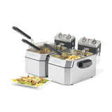 Two stainless steel electric deep fryers with black lids and control panels sit side by side. One fryer basket is lifted, showing golden fries inside. A white plate with fried food and garnishes sits in front of the countertop fryers.