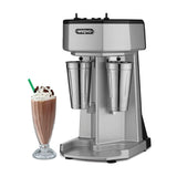 A Waring double spindle drink mixer with a sleek silver finish stands beside a glass of chocolate milkshake topped with whipped cream, chocolate shavings, and a green-striped straw. The drink mixer features two stainless steel mixing cups.