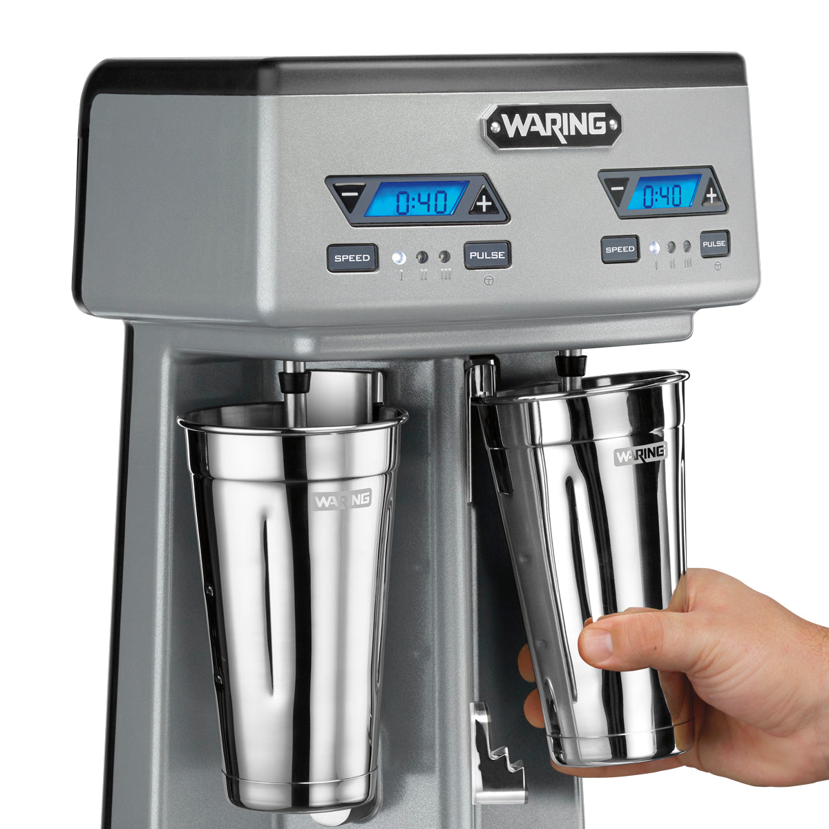 A close-up of a silver Waring Drink Mixer with a double spindle design and two stainless steel cups; one cup is being held by a hand. The digital display above each cup shows 0:40, with speed and pulse controls on the front panel.