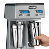 A close-up of a silver Waring Drink Mixer with a double spindle design and two stainless steel cups; one cup is being held by a hand. The digital display above each cup shows 0:40, with speed and pulse controls on the front panel.