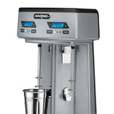 The Waring Double Spindle Drink Mixer features two mixing spindles, digital blue LED timers, control buttons for speed and pulse, and removable metal cups. The “Waring” brand label is displayed on the top front.