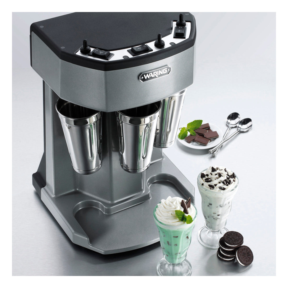 A silver triple spindle Drink Mixer with three metal cups sits on the counter. In front are two milkshakes—one mint chocolate and one cookies-and-cream—topped with whipped cream, surrounded by cookies, mint, and spoons.