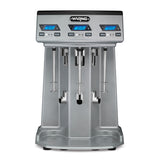The Waring WDM360TX Drink Mixer features a modern, industrial design with three Triple Spindle blenders, each with digital blue LED displays and control buttons above. The wide base includes grooves for placing cups under each spindle.