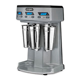 A commercial Waring WDM360TX Triple Spindle Drink Mixer with three stainless steel mixing cups, digital timers, blue LED displays, and black start/stop buttons on a sleek gray metallic base. The robust machine stands confidently on four small feet.