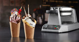 Two brown paper cups hold rolled waffles filled with ice cream and toppings. In the background, a Waring bubble waffle maker with non-stick plates hints at the delicious creations—chocolate, strawberry, cookies-and-cream scoops, sprinkles, and wafer sticks.
