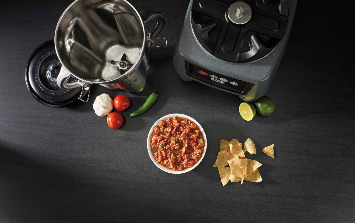 Overhead view of a heavy-duty blender with its lid off, two tomatoes, garlic, a jalapeño, halved lime, bowl of salsa, and tortilla chips on a dark countertop—perfect for preparing homemade salsa with fresh ingredients.
