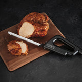 A black cordless carving knife labeled “Waring” rests on a wooden cutting board next to a partially sliced cooked roast. One thick slice and the remaining roast are visible on the dark textured surface.