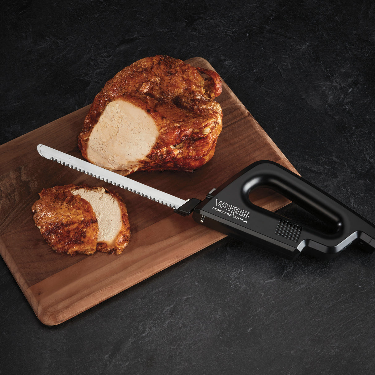 A cooked, seasoned boneless meat roast sits on a wooden cutting board. Two slices have been cut off. A Commercial electric carving knife labeled Waring, with an adjustable slicing guide, rests on the board, its serrated blade partly under a slice.