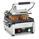 A stainless steel Compact Toasting Grill with a black handle is open, grilling two sandwiches. The sandwiches have golden brown bread and a visible filling of white cream and sliced strawberries. It features adjustable thermostats and displays the brand Waring.