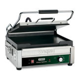 A stainless steel Waring commercial panini grill with a partially open ribbed top plate, flat cast iron cooking surface, black handle, side handles, and a front control panel featuring an adjustable thermostat and labeled Waring Tostato Supremo.