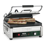 A stainless steel Waring Panini Grill with a cast iron cooking surface and partially open lid grills two long sandwiches. The control panel displays an adjustable thermostat and the label Waring Commercial Tostato Supremo. Fillings like meat and cheese are visible.